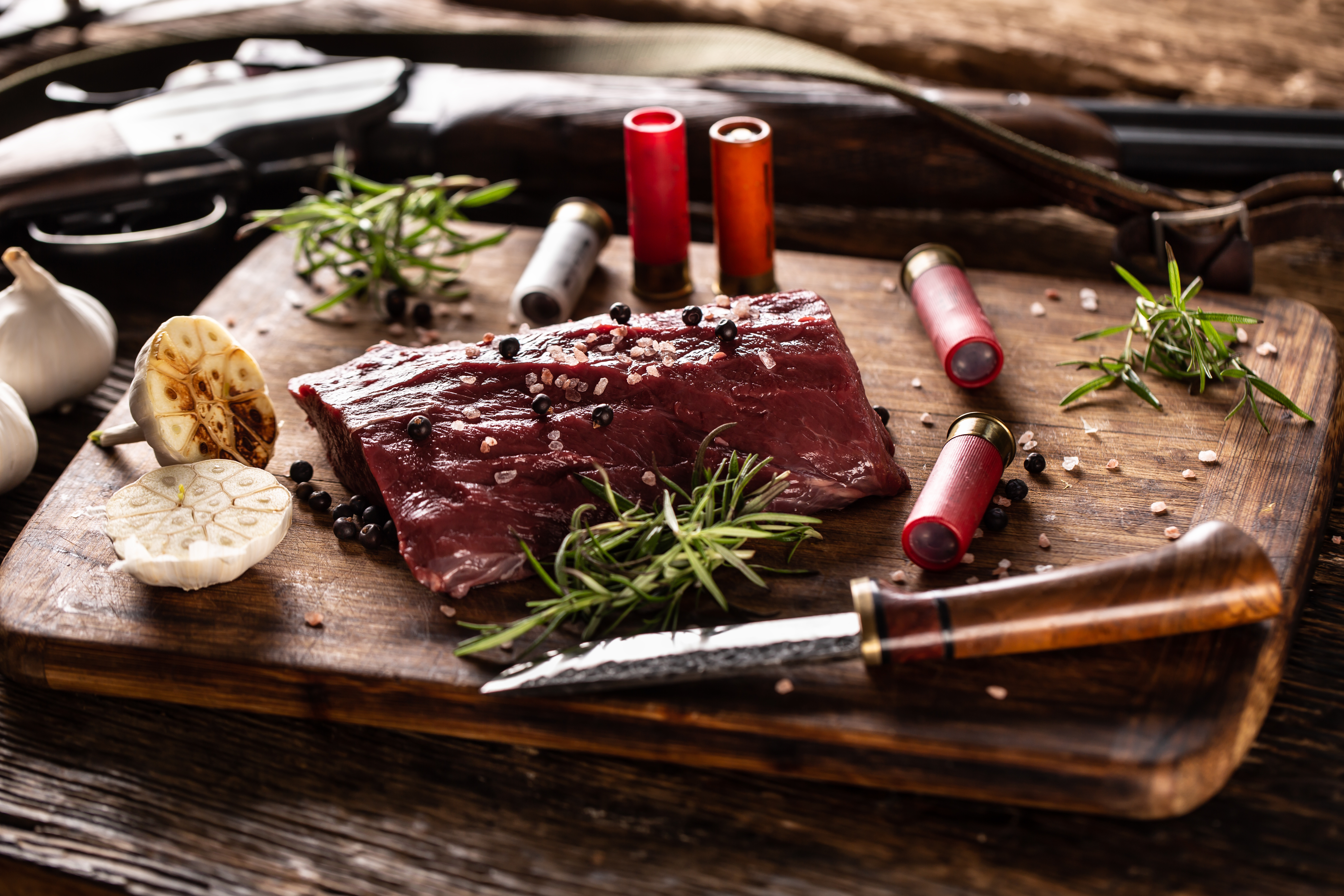 A raw venison loin on a wooden board with rosemary, garlic, a hunting knife, and shotgun shells.