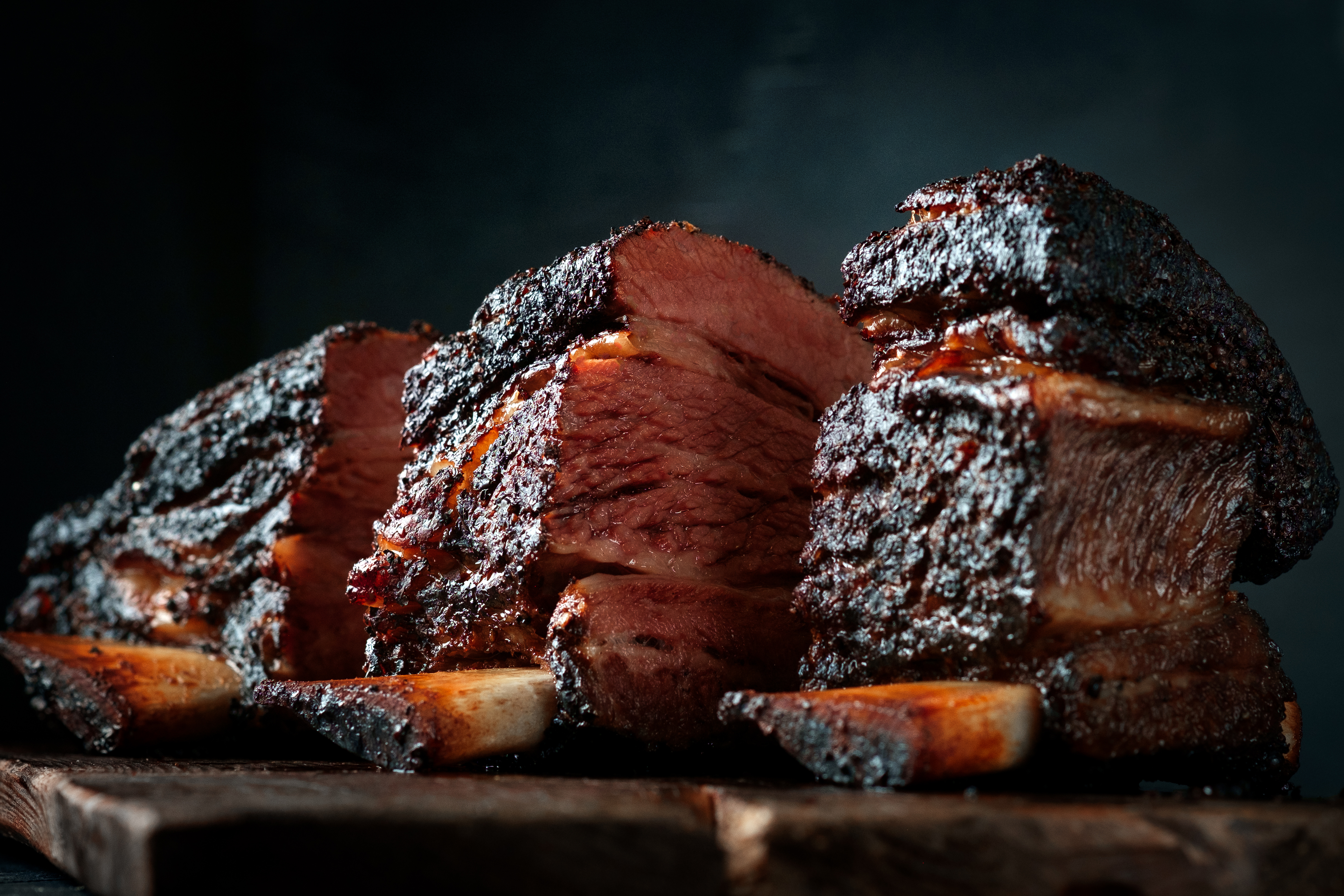 Three smoked beef ribs with dark bark on a wooden board, bone visible under the meat.