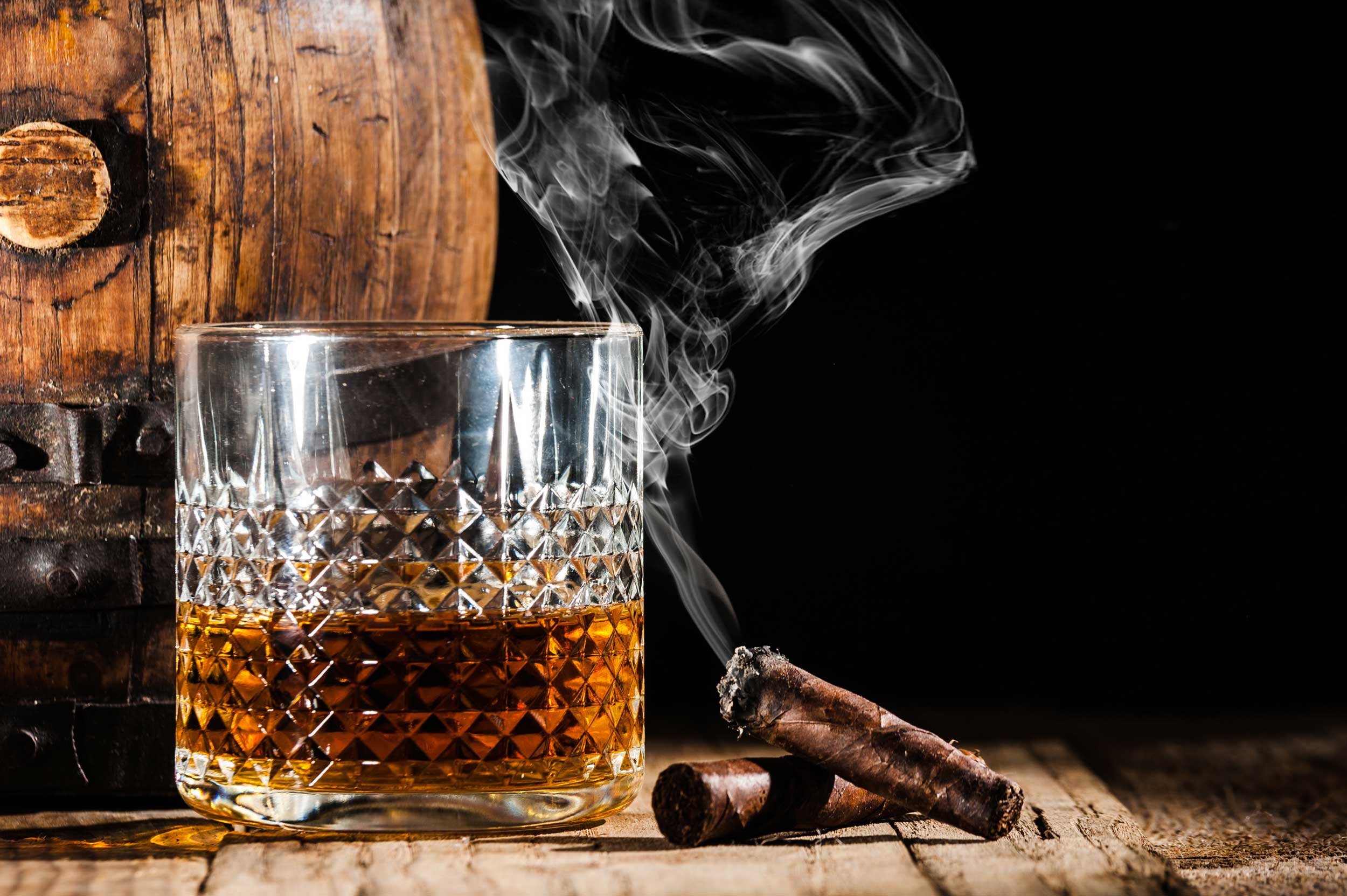 A cut crystal glass of amber spirit beside two cigars and a small oak barrel.
