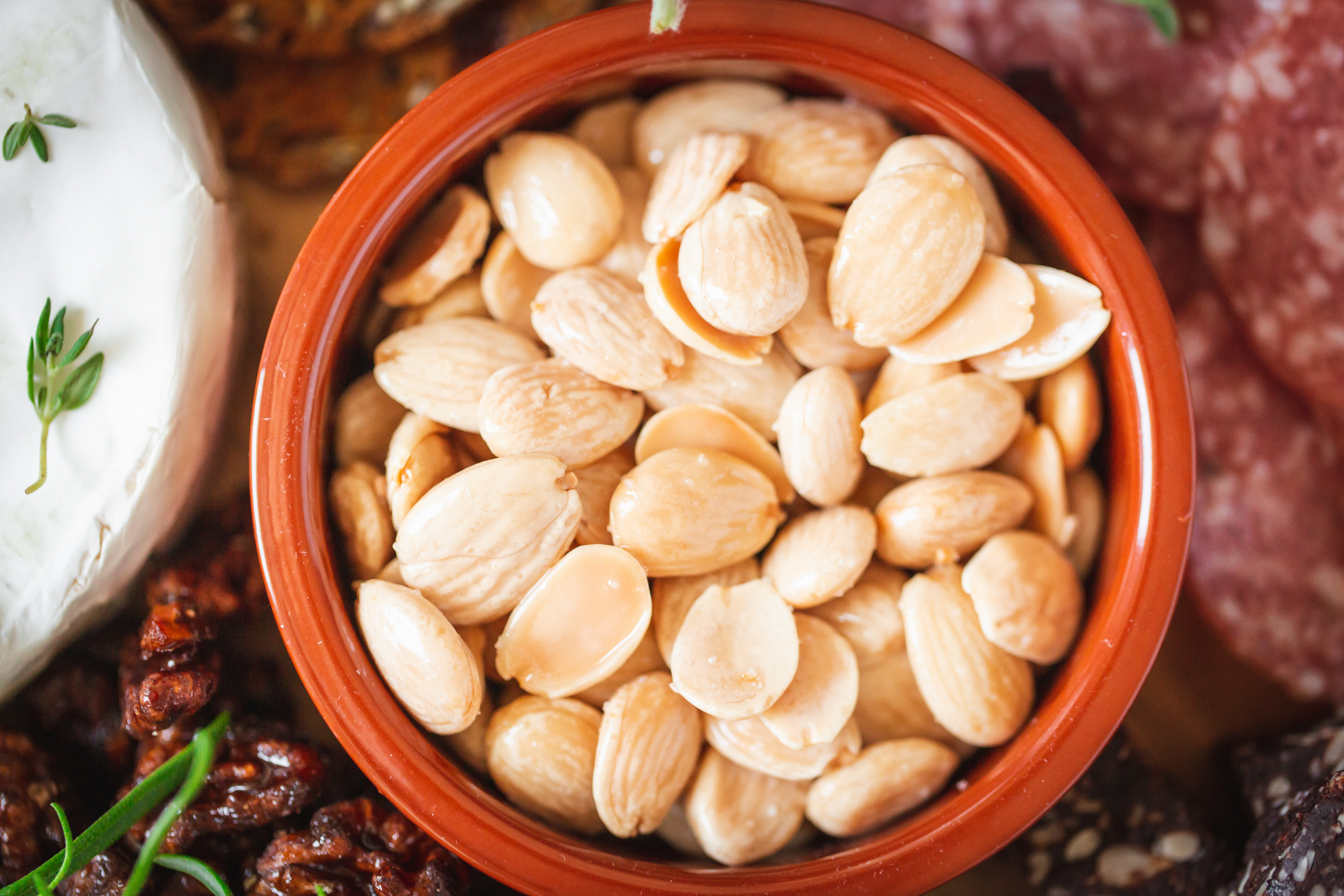 Blanched almonds in a terracotta bowl beside brie, walnuts, and sliced salami.