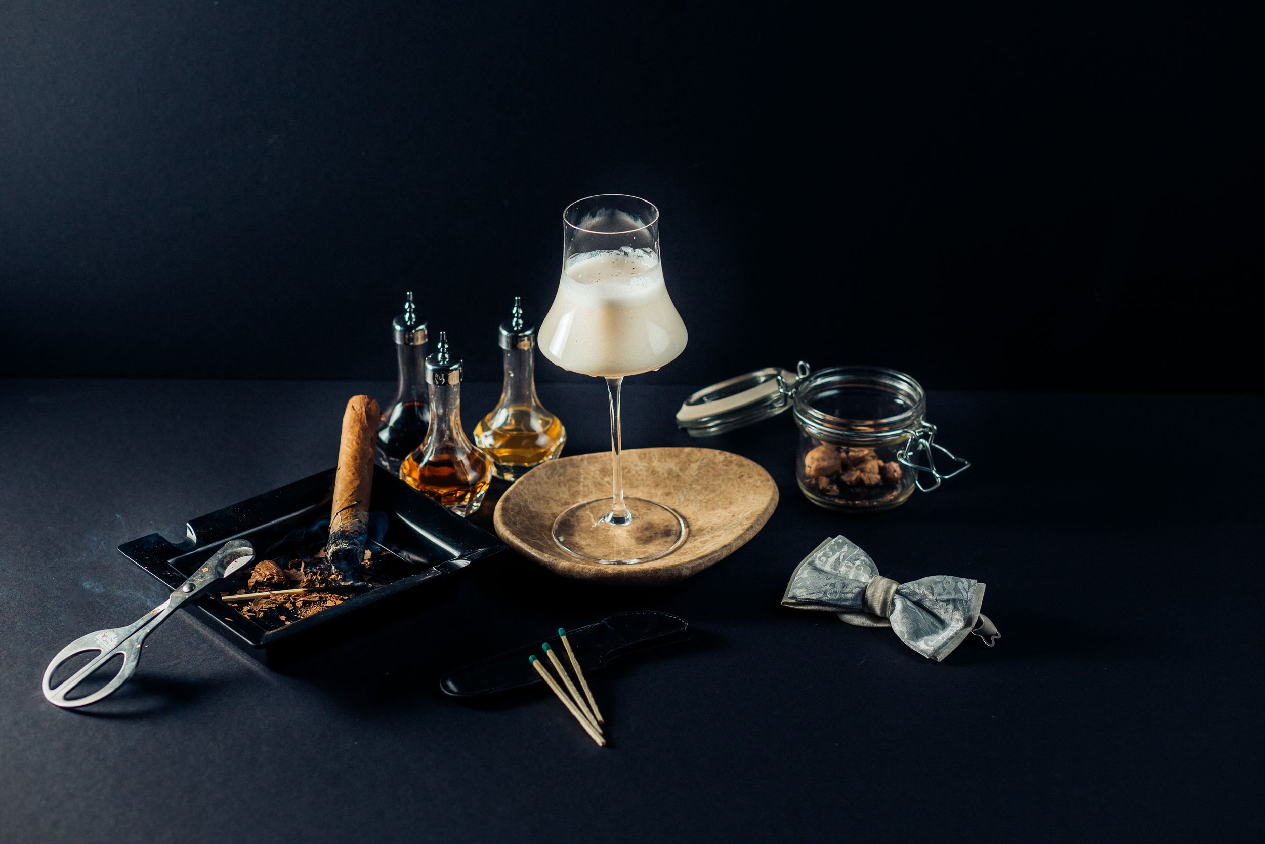 A jar of roasted almonds beside a lit cigar, cutter, and a frothed glass.