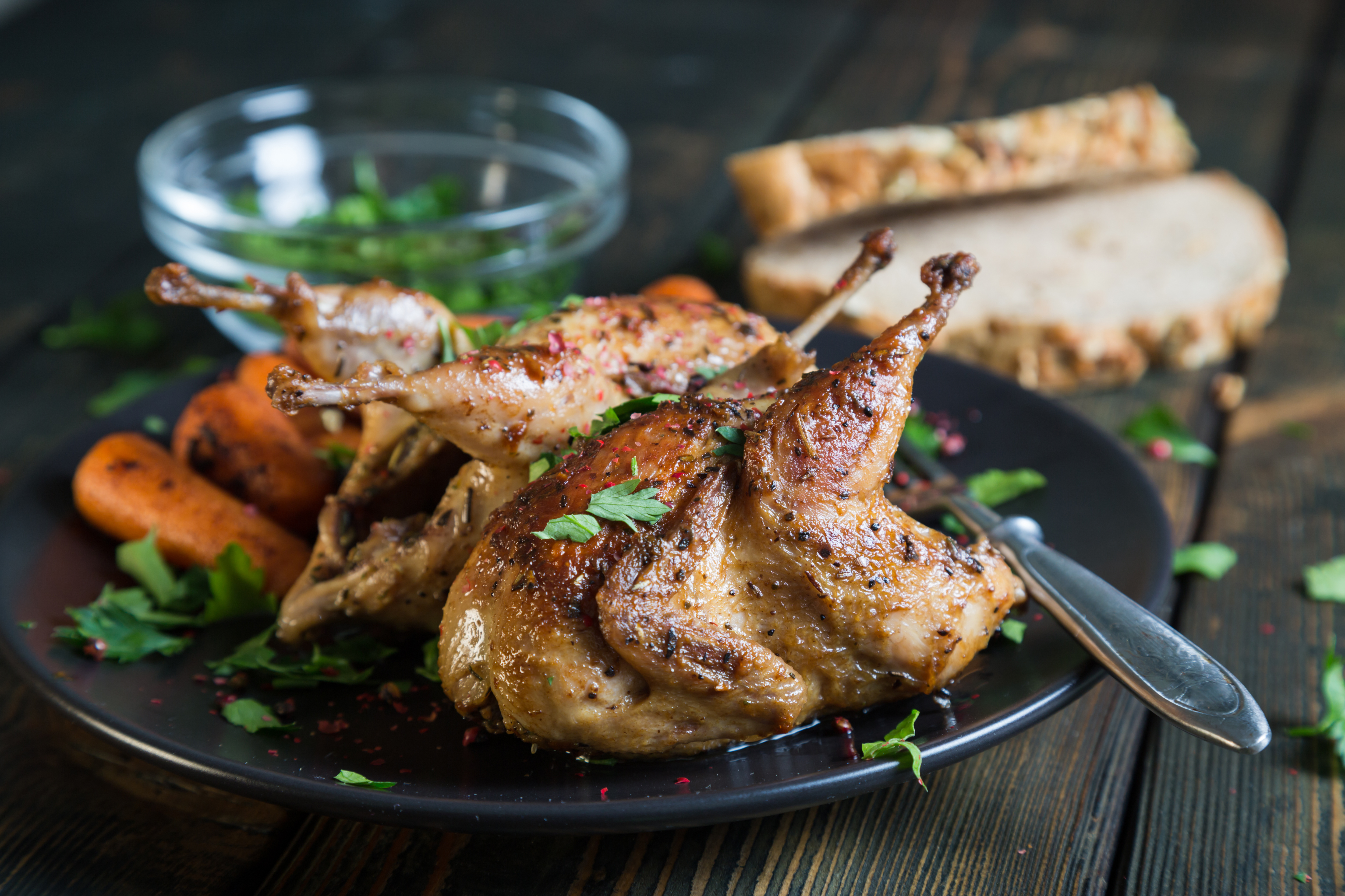 Two small roasted game birds on a dark plate with parsley, carrots, and red peppercorns.