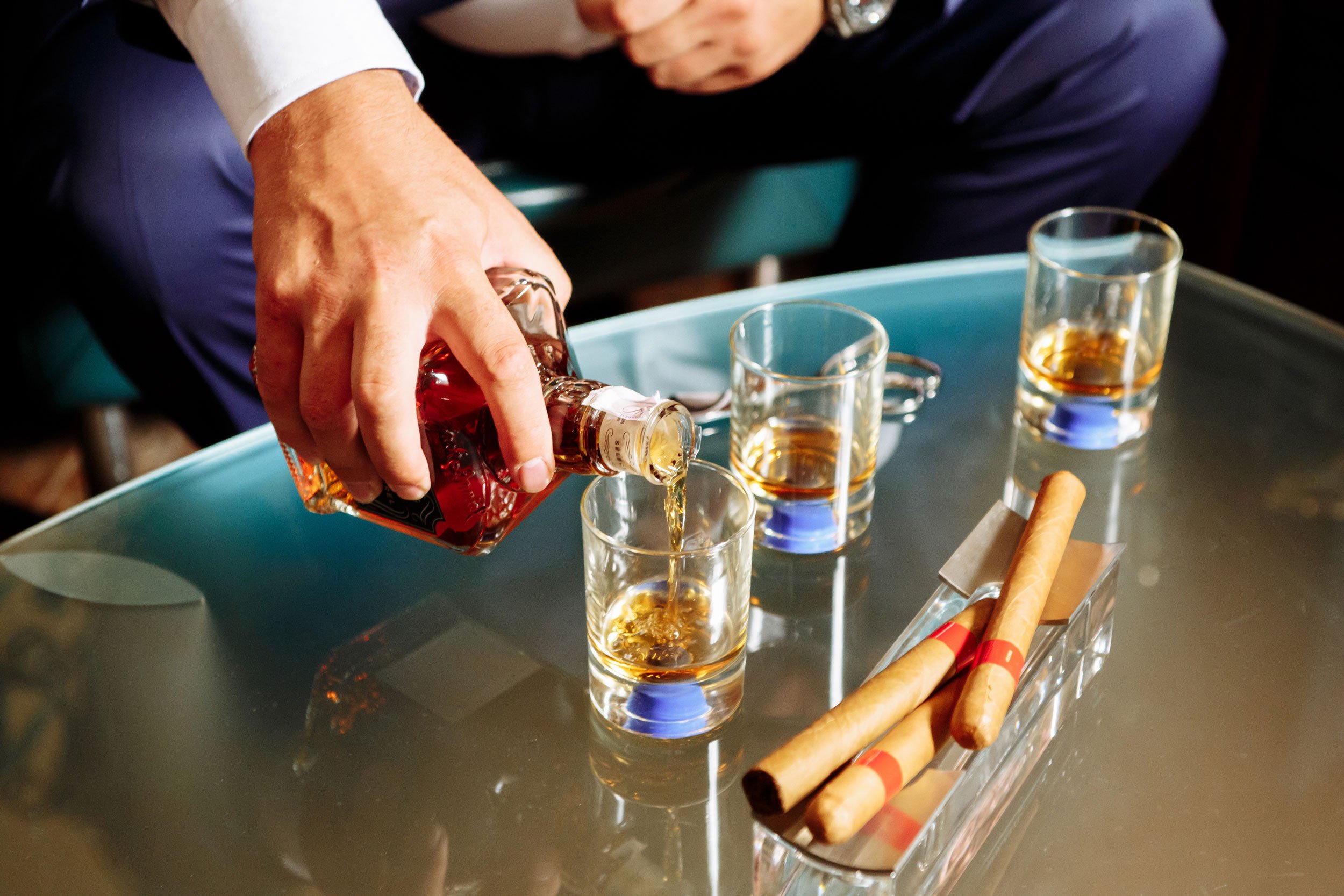Amber spirit poured into short glasses beside cigars in a glass ashtray.