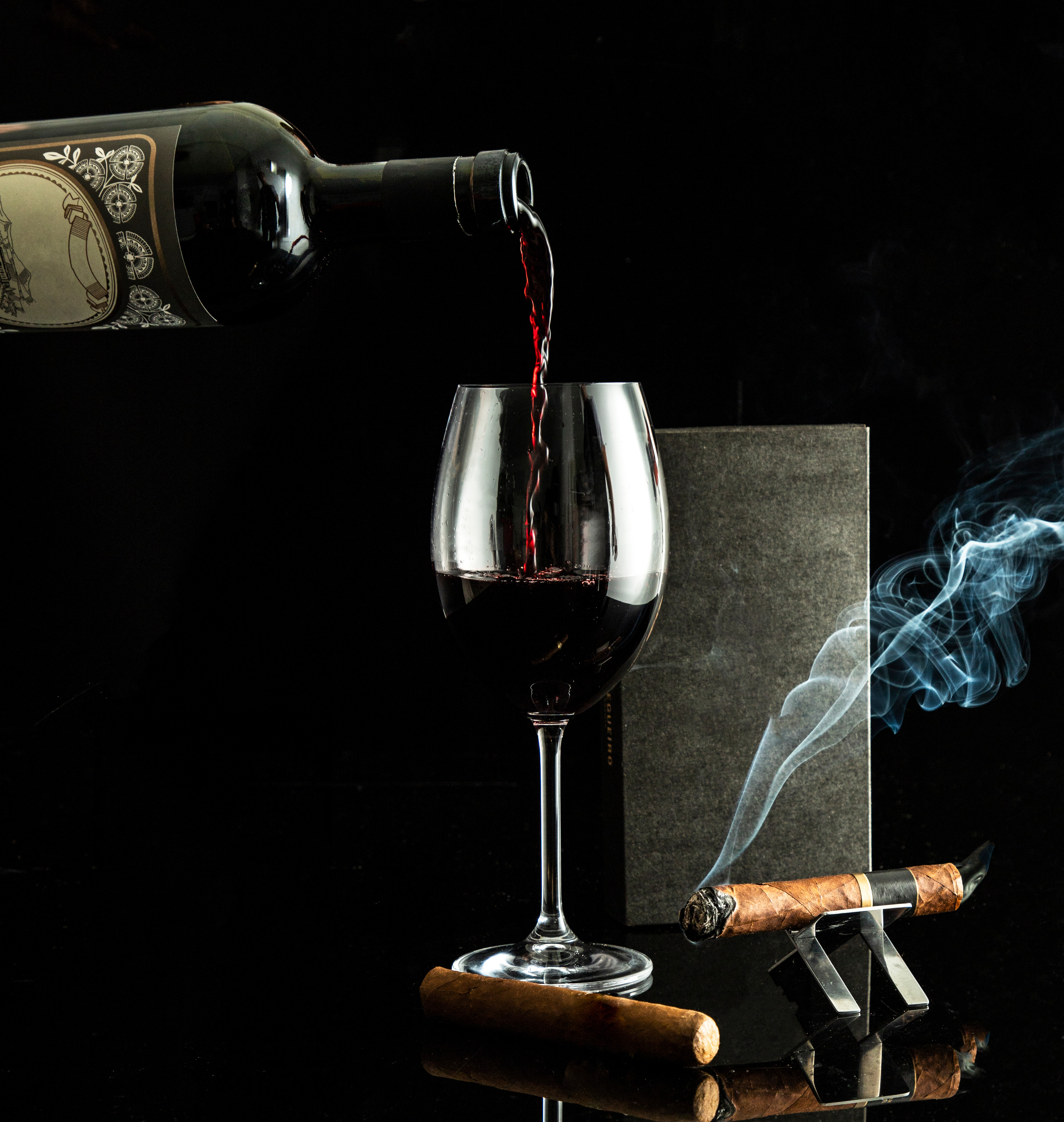 Red wine poured into a stemmed glass beside a lit cigar on a silver stand.